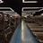 A spacious gym with multiple rows of treadmills and weightlifting benches on either side. The lighting features geometric patterns on the ceiling, creating a modern and organized ambiance. People are actively using the equipment, engaging in fitness activities.