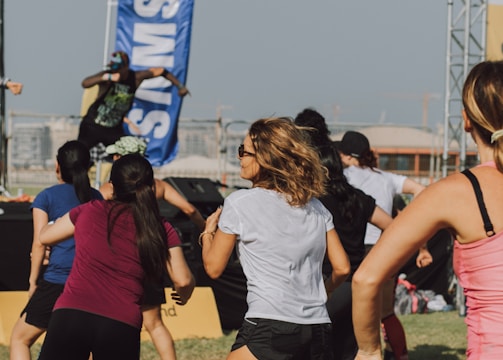 A group of people appears to be participating in an outdoor fitness class or dance session. They are dressed in athletic clothing, and a blue banner can be seen in the background, along with a stage. The setting suggests a lively and energetic atmosphere.