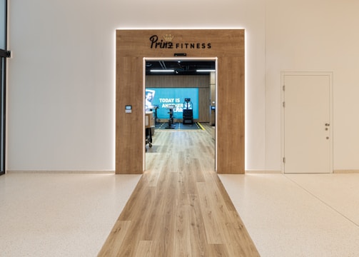 A modern fitness center entrance with wooden framing and the sign 'PRIME FITNESS' above the doorway. The interior features a spacious gym area with workout equipment visible and a motivational banner on the back wall. The flooring transitions from glossy tiles to a wood finish leading into the facility.