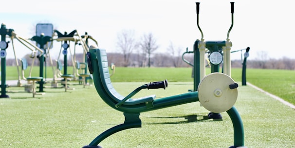 An outdoor gym with several pieces of exercise equipment arranged on a green artificial turf. The image focuses on a leg press machine in the foreground with other fitness machines visible in the background. The area is surrounded by an open, empty landscape.